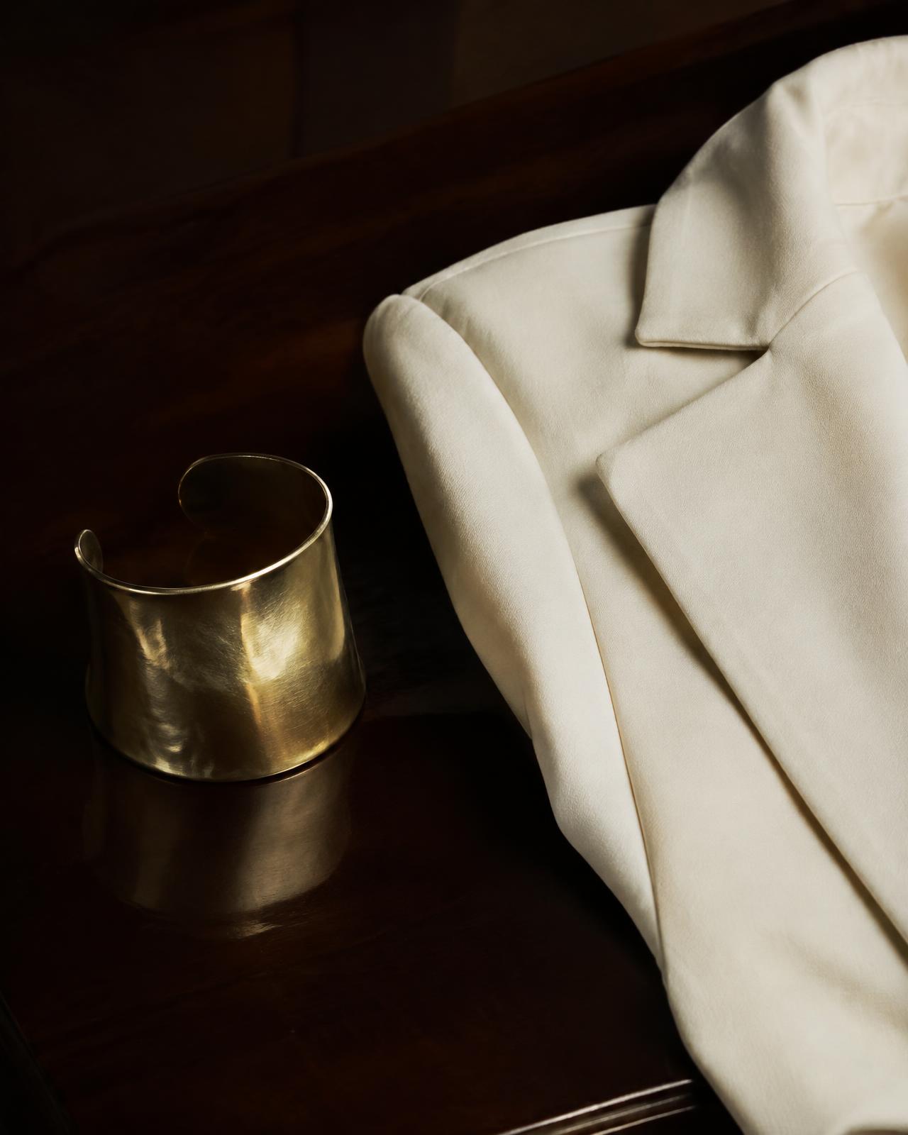 A sculptural brass cuff beside an ivory tailored jacket on dark lacquered wood
